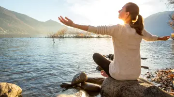 Junge Frau sitzt mit ausgebreiteten Armen am Ufer eines Sees und genießt die Sonne.